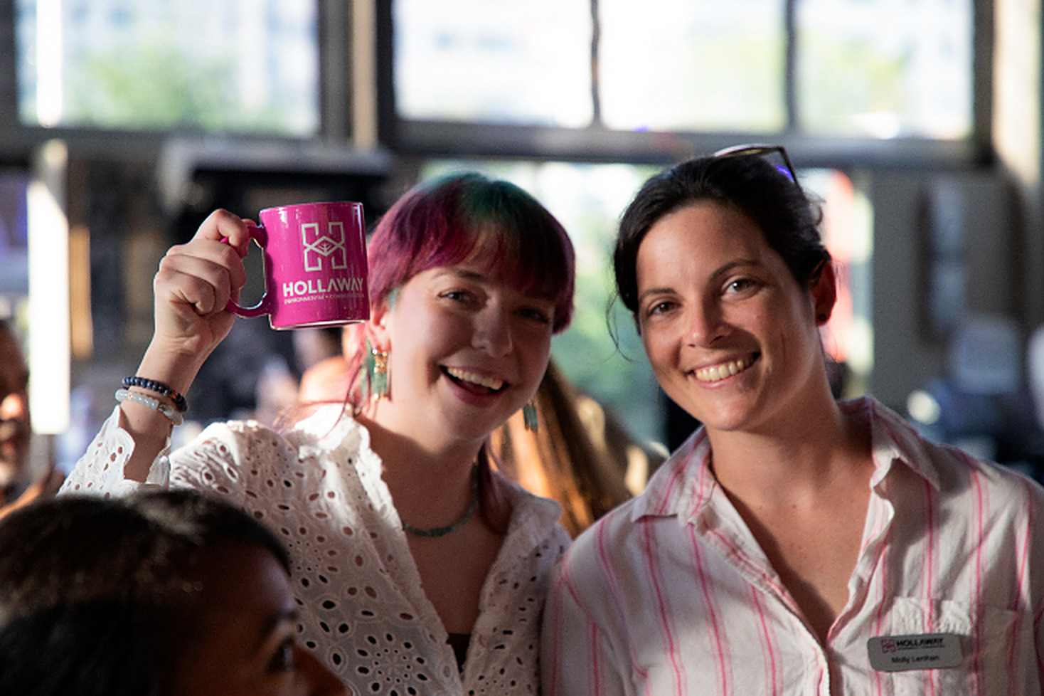 Two Hollaway team members smiling, one raising a pink Hollaway mug